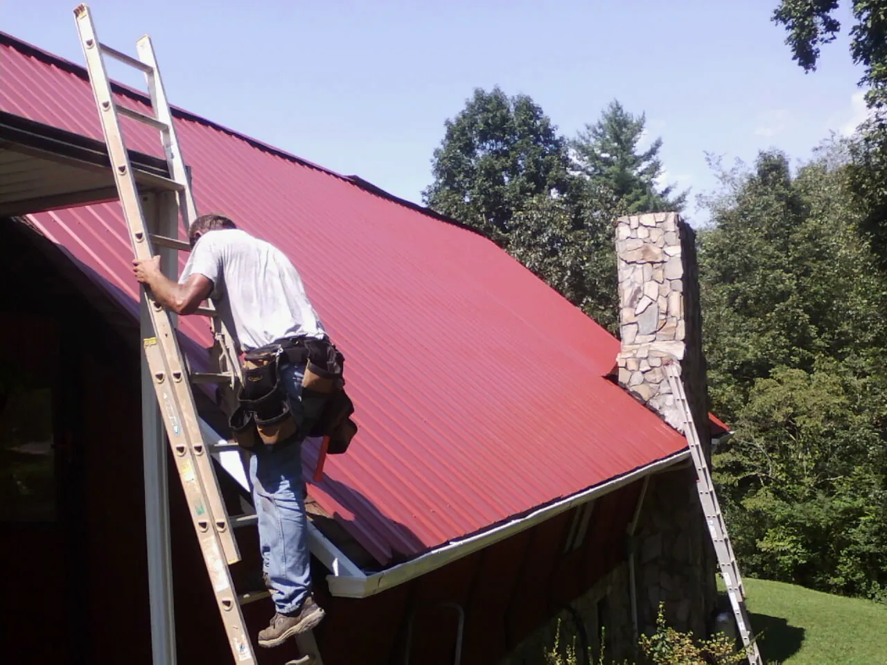 Roofer on a ladder beside a red metal roof and stone chimney — Soffit & Fascia Repair by Roofing Brothers in Conowingo